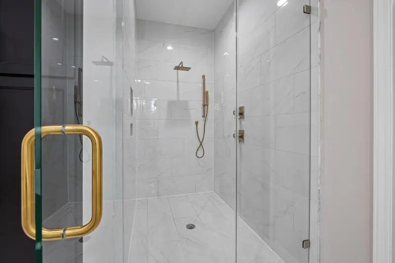 Walk-in shower with frameless clear glass, white marble-look tile and a brass door handle