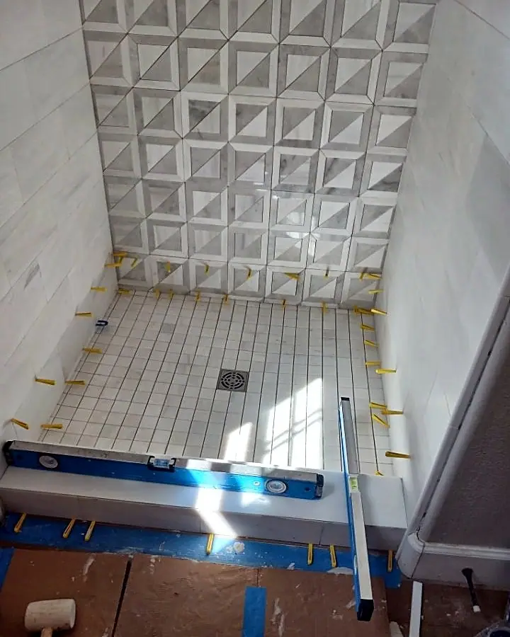 New shower floor tile being set with leveling clips, below a wall of white dimensional tile